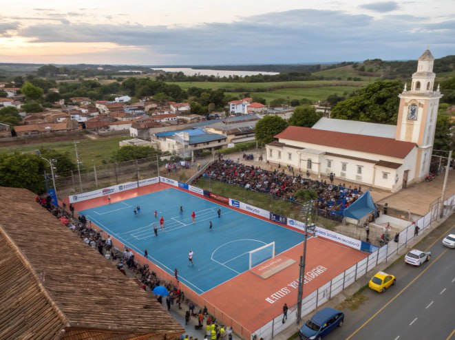 Quadra de futsal em cidade do Paraná com igreja histórica ao lado e arquibancadas cheias durante partida.