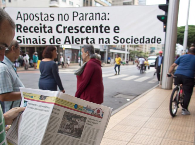 Faixa em rua de Curitiba destaca apostas no Paraná com receita crescente e alertas sociais, enquanto pessoas circulam e um homem lê jornal.